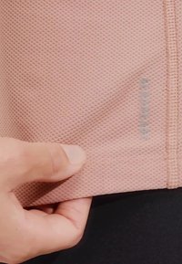 Lichtroze gestructureerde stof met een diamanten patroon en een gaasstructuur, met een label waarop "AEROREADY" in het grijs staat. Hand zichtbaar.