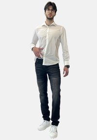 Camisa blanca de manga larga con botones, jeans oscuros con rayas sutiles y zapatillas blancas. Persona de pie en posición neutral.