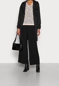 Schwarzer Trenchcoat über einem hell gemusterten Oberteil, kombiniert mit weit geschnittenen schwarzen Hosen und schwarzen Stiefeletten. Als Accessoires dient eine strukturierte schwarze Handtasche.