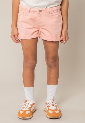 Persoon die lichtroze opgerolde korte broek, witte geribbelde sokken en oranje-witte sneakers draagt, staand op een effen achtergrond.
