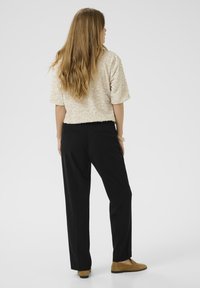 Bluză cu mânecă scurtă, texturată, de culoare bej, cu paiete, asortată cu pantaloni negri, croiți, și pantofi loafer maro deschis, văzută din spate.