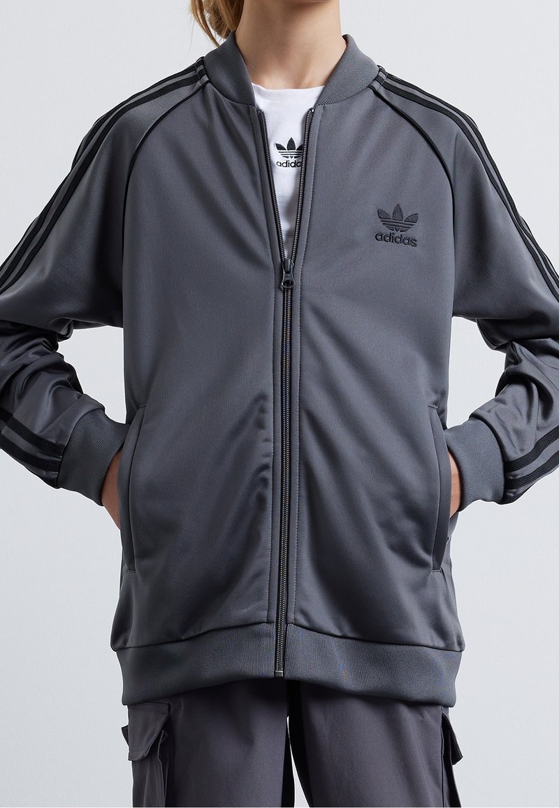 Grå träningsjacka med en mjuk textur, blixtlås fram, ribbade manchetter och tre svarta ränder på ärmarna; broderad Adidas-logotyp på bröstet.