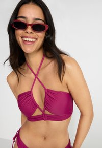 Magenta bikini viršūnė su rinkta tekstūra ir kryžminėmis petnešėlėmis. Suderinta su atitinkamomis kelnaitėmis, modelis nešioja tamsiai rožinius ovalius akinius nuo saulės.