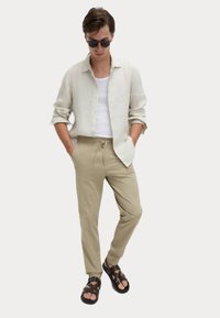 Linnen overhemd met knoopsluiting in lichtbeige, witte geribde tanktop, beige broek met trekkoorden en bruin sandalen met bandjes. Soepele textuur overal.