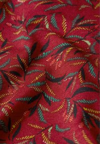 Punainen kangas, jossa on teksturoitu kudonta, esittelee värikästä lehtikuviota teal, keltainen, musta ja vaaleanvihreä, ja taitteet luovat syvyyttä.