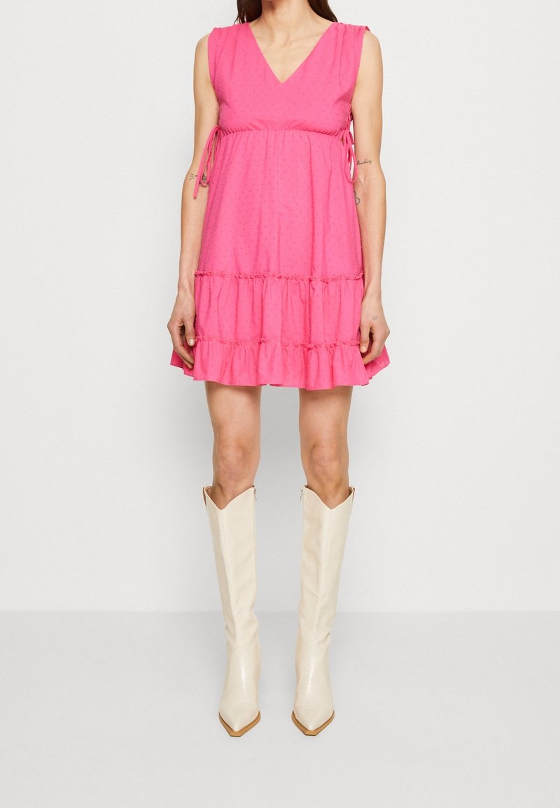 Femme portant une robe rose sans manches à encolure en V avec des volants, assortie à des bottes blanches en cuir montant jusqu'aux genoux, debout devant un fond blanc.