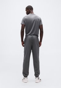 Ά νδρας που στέκεται πλάτη φορώντας γκρι αθλητικό παντελόνι και ταιριαστό κοντομάνικο πουκάμισο, με άσπρα και μαύρα αθλητικά παπούτσια σε λευκό φόντο.