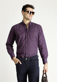 Camicia a maniche lunghe a quadri rossi e blu con colletto button-down e taschino, abbinata a jeans scuri e occhiali da sole.