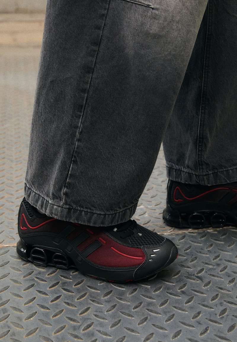 Sorte og røde treningssko med mesh-struktur, buet gummisåle og distinkte utskjæringsdetaljer, parret med grå denim-bukser.