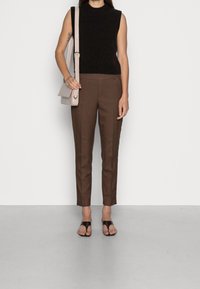 Svart ärmlös stickad toppen, bruna skräddarsydda byxor och svarta sandaler. Ljusgrå axelremsväska med rem. Platt, slät yta som bakgrund.