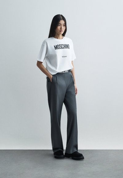 Femeie în picioare cu mâinile în buzunare, purtând un tricou alb Moschino Milano, pantaloni gri și pantofi negri, pe un fundal deschis, simplu.