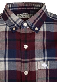 Koszula w kratę w kolorach śliwkowym, granatowym i szarym, z kołnierzykiem w szpic, dwoma guzikami oraz małym białym haftowanym logo na kieszeni.