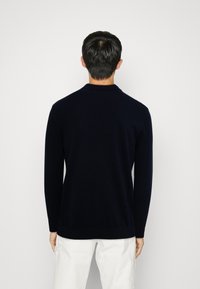 Man som vänder bort mot kameran, iförd en marinblå långärmad tröja och vita byxor, mot en enkel ljus bakgrund.