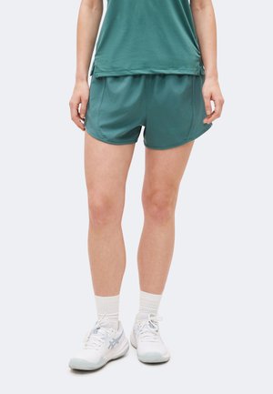 Osoba ubrana w turkusowe sportowe spodenki i top, białe skarpetki o długości za kostkę oraz białe buty do biegania, stojąca na tle jednolitego tła.