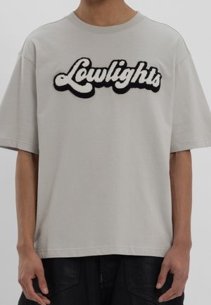 Jasnoszary bawełniany t-shirt z teksturowanym, czarno-białym logo „Lowlights” na przodzie, o luźnym kroju i krótkich rękawach.