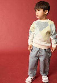 Un pull clair avec un motif géométrique en jaune, gris et bleu ; associé à un pantalon gris et des baskets blanches. Fond rouge.