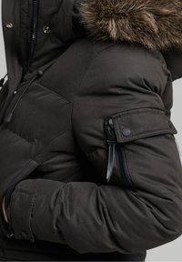 Chaqueta de invierno negra con capucha con ribete de piel, bolsillo en el brazo con cremallera, solapa con botones y manga acolchada, llevada con los brazos cruzados.