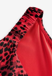 Crvena kupaćica s crnim leopard uzorcima; teksturirana, rastezljiva tkanina; u njoj se ističu nabranosti duž izreza i naramenica.