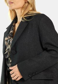 Abrigo negro con patrón de espiga y acabado texturizado, que presenta un diseño de doble botonadura y botones grandes, además de un clásico cuello de solapa.