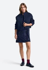 Námornícky modrý skrátený kabátik s gombíkmi, doplnený o námornícky modré šaty s flitrovanými kvetinovými vzormi. Modelka má na sebe bordové ponožky a čierne mokasíny.