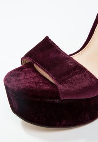 Gros plan d'une sandale plateforme en velours violet foncé avec une large bride avant et une doublure intérieure beige.