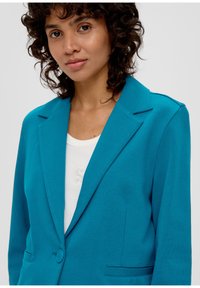 Turkusowa marynarka zapinana na jeden guzik, z klapami i dwiema przednimi kieszeniami, zestawiona z białą, bezrękawnikową bluzką.