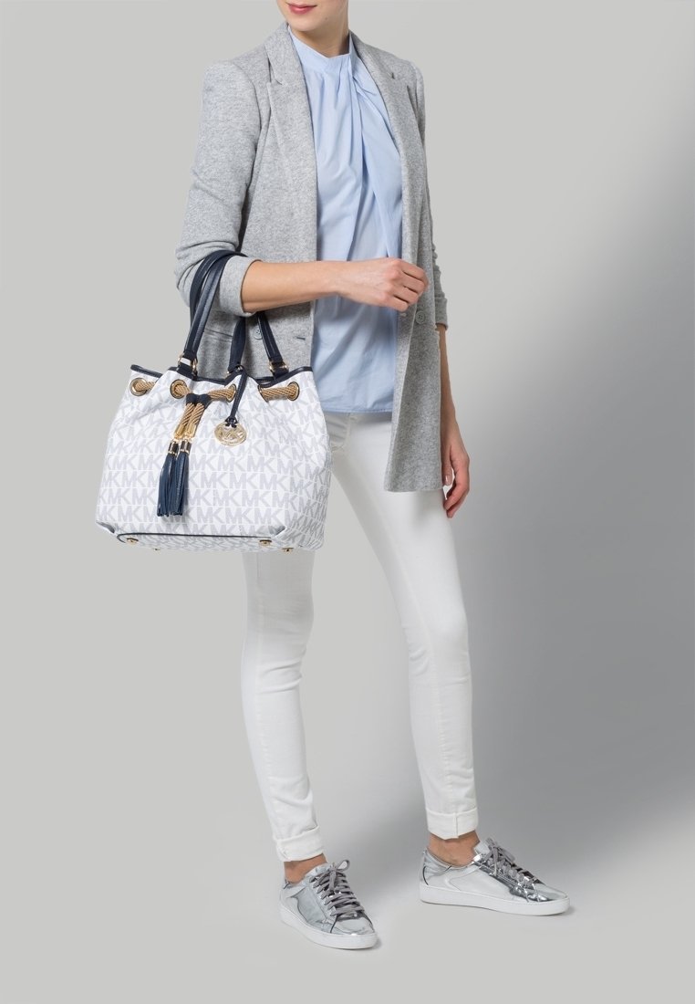 Femme portant un chemisier bleu clair, un blazer gris, un pantalon blanc, des baskets argentées, tenant un sac à main de créateur blanc avec des accents bleu marine et or.