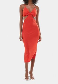 Červené žebrované šaty se špagetovými ramínky, s uzlem na přední části a výřezy na stranách. Délka do půli lýtek se mírně asymetrickým lemováním.