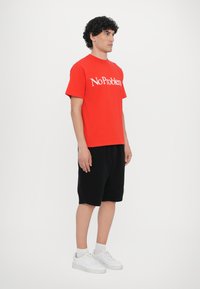 Rotes Kurzarm-T-Shirt mit "NoProblem" in Weiß bedruckt. Kombiniert mit schwarzen Shorts und weißen Sneakers. Glattes Material, lässige Passform.