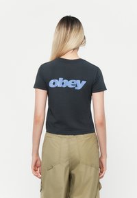 Black cotton t-shirt with short sleeves, featuring light blue "obey" text on the back. Paired with light khaki cargo pants.