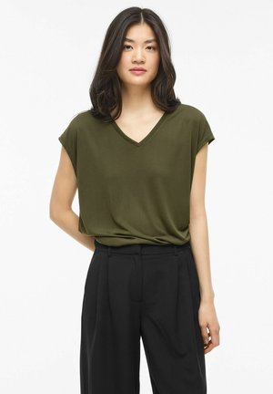 Mujer con cabello oscuro hasta los hombros, vistiendo una blusa verde oliva de manga corta con escote en V y pantalones negros plisados de cintura alta, de pie contra un fondo blanco.