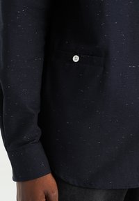 Camicia blu con bottonatura frontale e tasca, caratterizzata da una texture maculata e da un bottone bianco. Il tessuto appare durevole e ben strutturato.