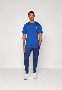 Modra atletska majica z belim logotipom, kombinirana z mornarsko modrimi hlačami z belimi stranskimi črtami in teksturirano tkanino, obuti z belimi športnimi copati.