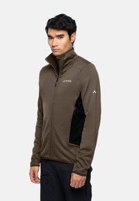 Braune Zip-Jacke mit hohem Kragen, strukturiertem Stoff und schwarzen Seitentaschen. Mit einem Logo auf der linken Brust und Akzentdetails.