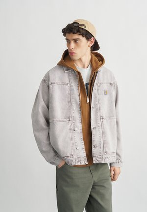 Giovane uomo con berretto beige indossato al contrario, felpa marrone con zip, giacca di jeans grigia e pantaloni verde oliva, in piedi con le mani parzialmente nelle tasche.