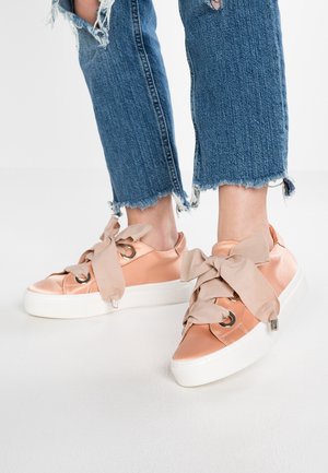 Nogi w znoszonych niebieskich dżinsach noszące błyszczące różowe satynowe sneakersy na platformie z dużymi beżowymi kokardami na białym tle.