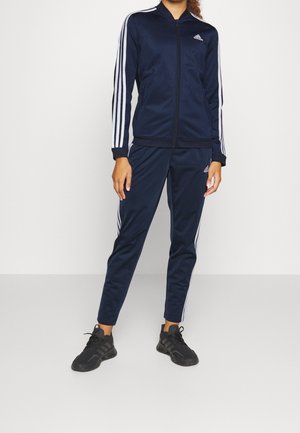 Personne portant un survêtement Adidas bleu marine avec des bandes blanches et des chaussures de sport noires, debout devant un fond uni clair.