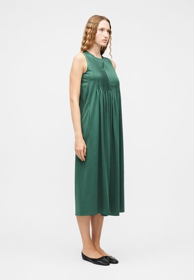 Γυναίκα που στέκεται φορώντας ένα αμάνικο, έως το γόνατο, σκούρο πράσινο πλισέ φόρεμα και μαύρα επίπεδα παπούτσια, πάνω σε απλό λευκό φόντο.