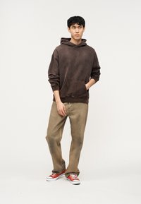 Νέος άνδρας που στέκεται, φοράει σκουρόκαφέ φούτερ με κουκούλα, ανοιχτό καφέ χαλαρά τζιν και κόκκινα χαμηλά αθλητικά παπούτσια σε λευκό φόντο.