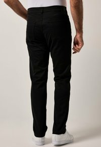 Pantalones slim-fit negros con una textura suave, bolsillos traseros y costuras en contraste, combinados con zapatillas blancas sobre un fondo claro.
