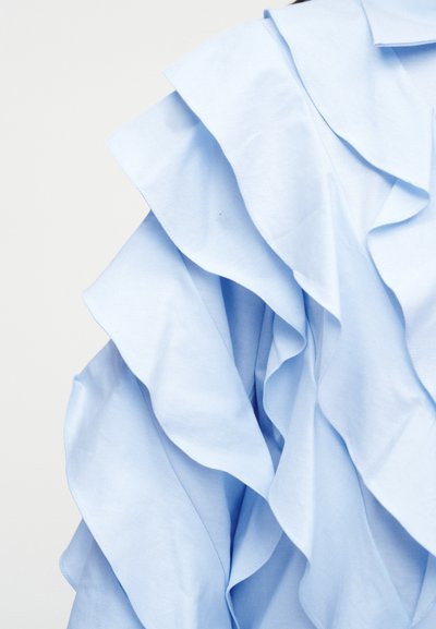Ανοιχτό μπλε ύφασμα με στρωματοποιημένες, κυματιστές βολάν που δημιουργούν απαλή, ογκώδη υφή πάνω σε απλό φόντο.