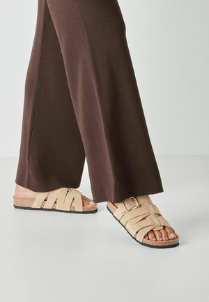 Sandales en daim beige avec des brides croisées et une boucle latérale, sur une semelle noire texturée, assorties à un pantalon large marron.