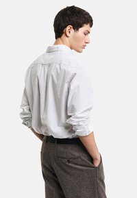 Camisa blanca de manga larga con pequeños lunares azules, cuello y mangas enrolladas; combinada con pantalones grises texturizados y cinturón negro.