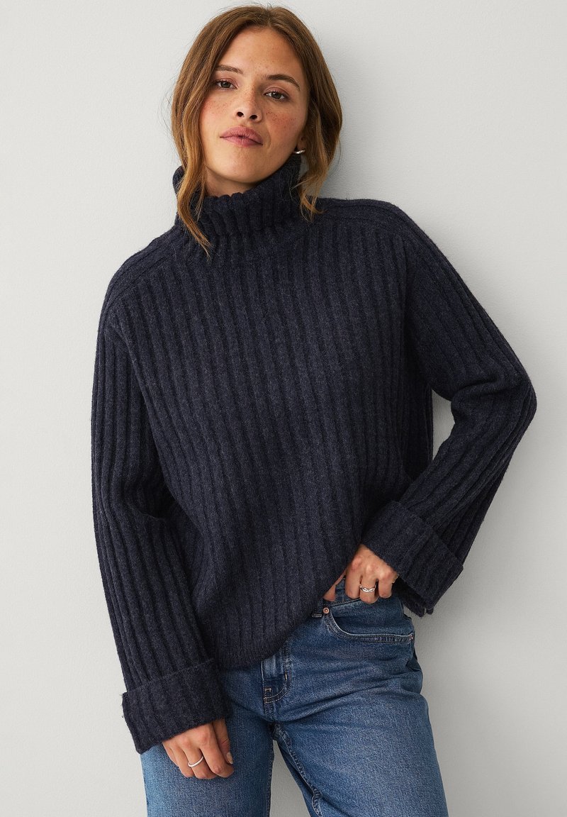 Navy ribbed turtleneck sweater with long sleeves, wide cuffs, and a textured fabric. Styled with blue denim jeans.