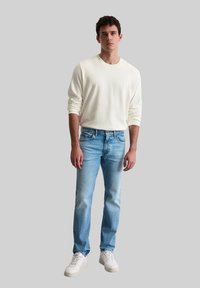 Chemise beige clair à manches longues, jeans bleus délavés et baskets blanches. La coupe est décontractée, avec un design relax et des détails de couture standards.