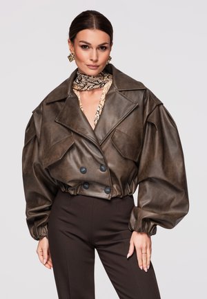 Femme portant une veste en cuir marron foncé surdimensionnée avec des poignets froncés, une écharpe à imprimé serpent et un pantalon noir taille haute, sur un fond neutre.