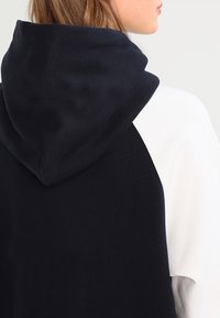 Kapuzensweatshirt mit gespaltenem Design: navyblauem Kapuzen- und Ärmeldesign, weißem Körper; weicher Stoff, glatte Textur und sichtbare Nähte.