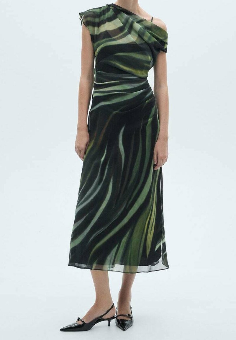 Robe maxi sans manches en tissu vert transparent à motif de feuilles, avec drapé et encolure asymétrique. Portée avec des chaussures noires à bouts pointus.