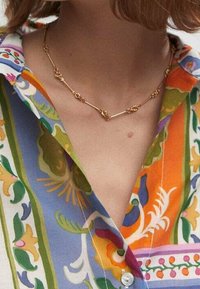 Gouden metalen ketting met kleine decoratieve clusters, gedragen over een meerkleurige, gestreepte blouse met bloemmotieven en levendige kleuren.