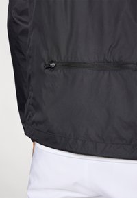 Melna viegla jaka ar gludu tekstūru, ar rāvējslēdzēja kabatu un pieguļošu apakšmalu. Apvienota ar baltām biksēm.